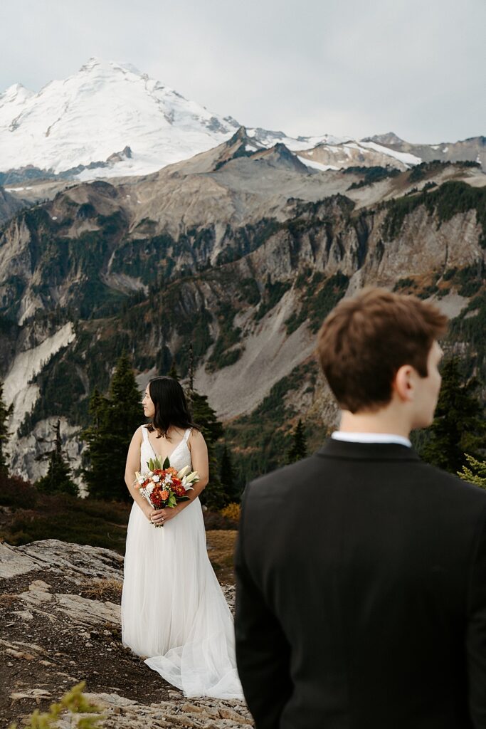 Groom in the immediate foreground looking to the right while bride is the main focus looking to the left and Mount Baker is behind her. 