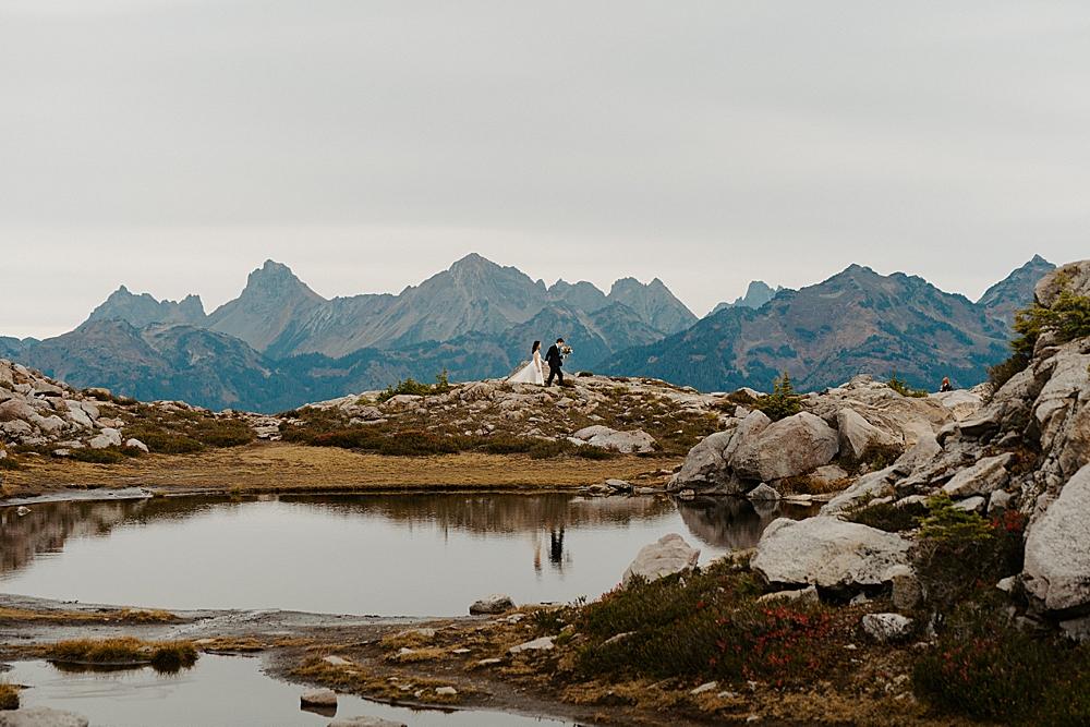 Bride and groom walking on a trail with Washington mountains behind them and their reflections showing in a small body of water in the foreground. 