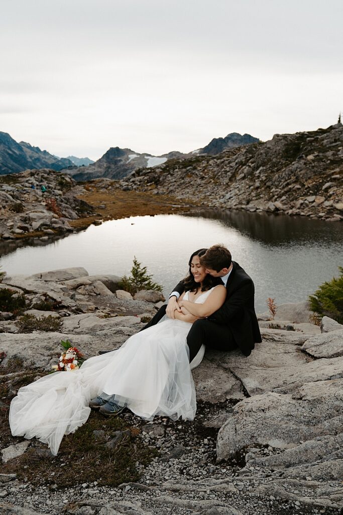 Mount Baker-Snoqualmie National Forest elopement photo of bride and groom sitting on a rock with a small body of water behind them.