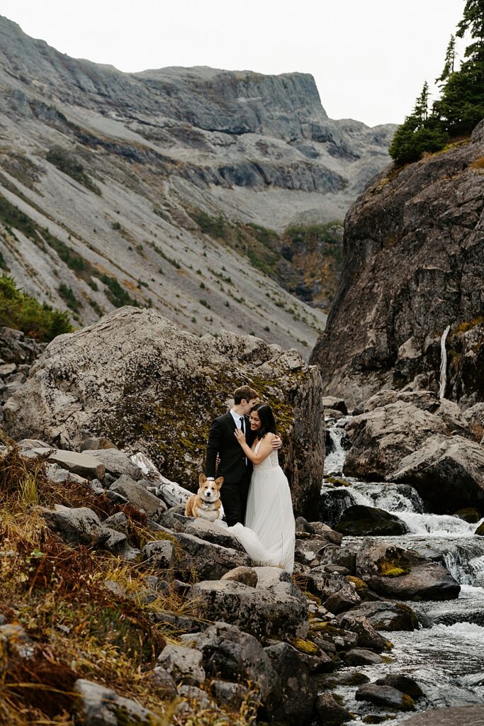 Bride, groom, and corgi standing on rocks by a creek for their Artist Point sunrise elopement. 