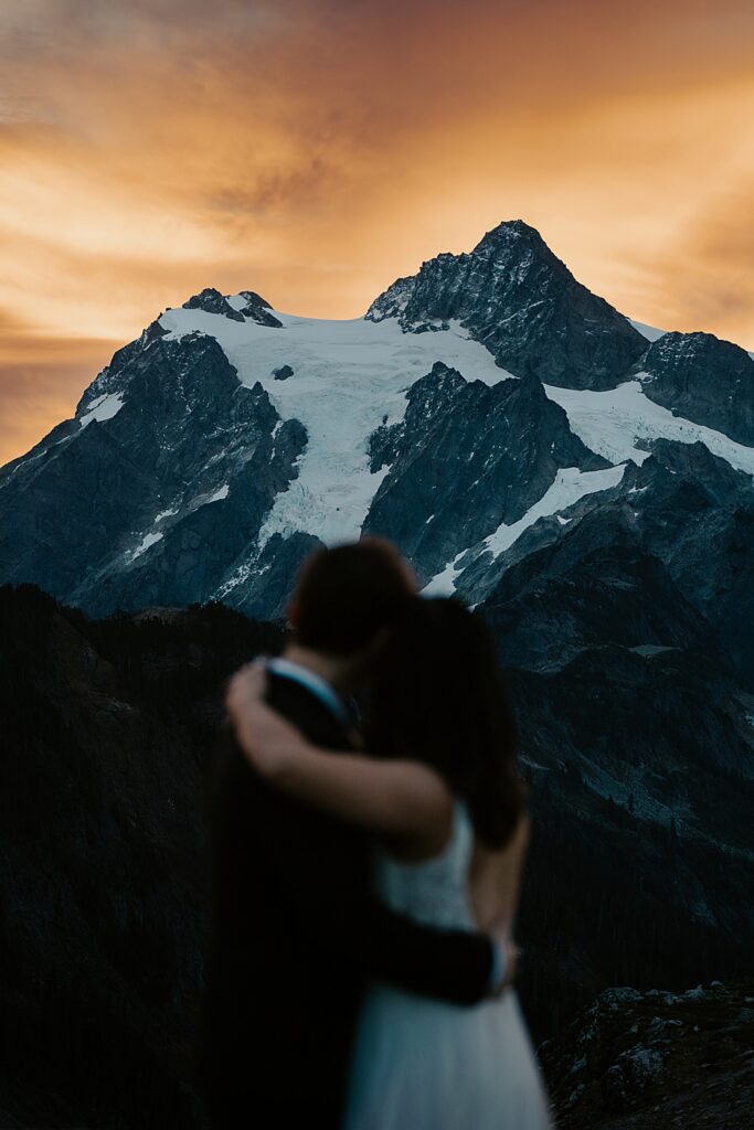 Blurry image of a bride and groom embracing and looking away with Mount Shuksan and sunrise in focus.