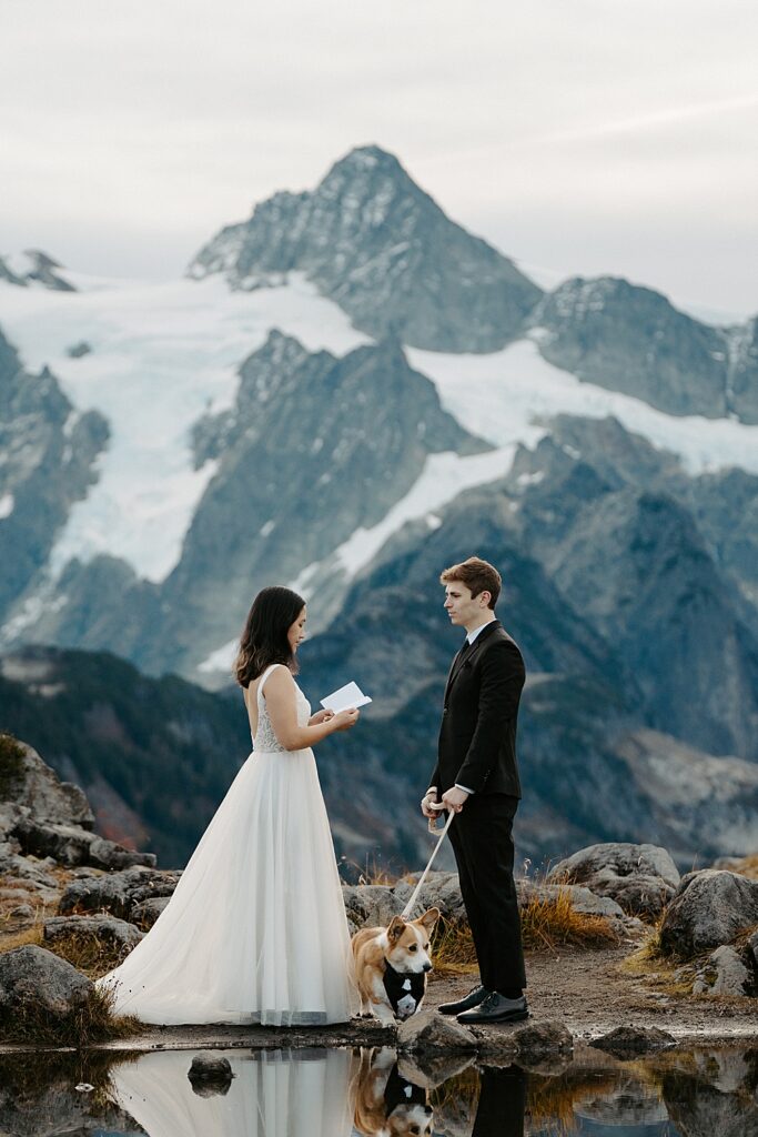 Bride saying elopement vows at Artist Point to groom who is holding a leashed corgi wearing a tuxedo collar.