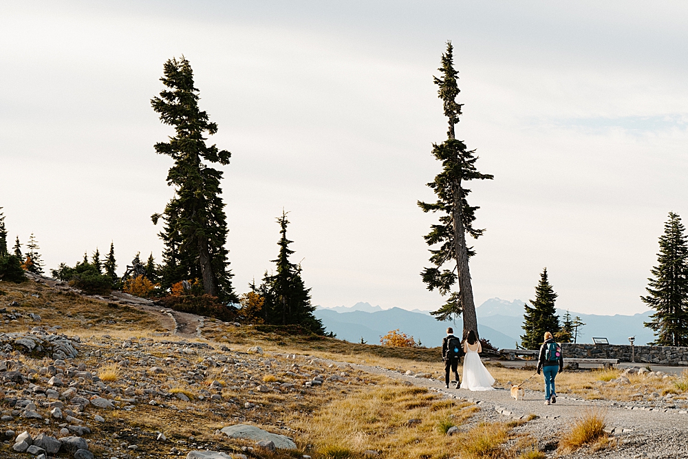 Bride and groom walking along a trail with a dog-handler and their corgi behind them.