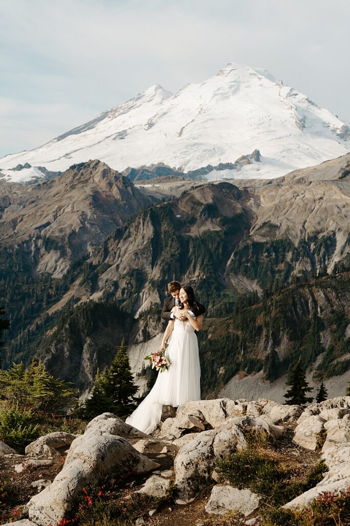 Bride and groom holding each other while standing on a rock with Mount Baker in the background.