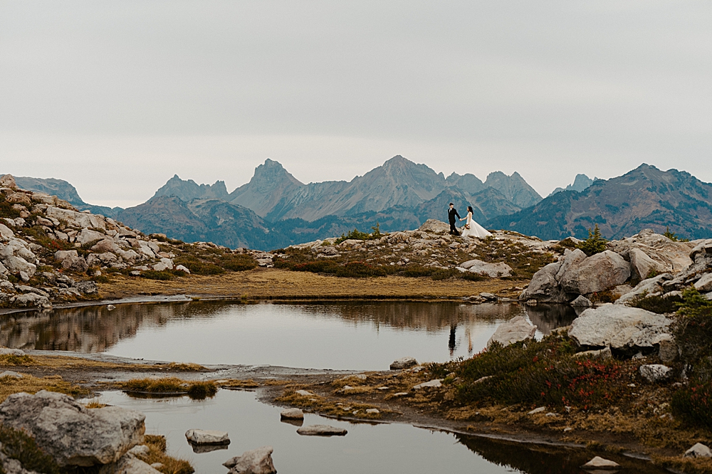 Bride and groom holding hands and walking on a trail with the mountains and a reflective pond up at Artist Point.