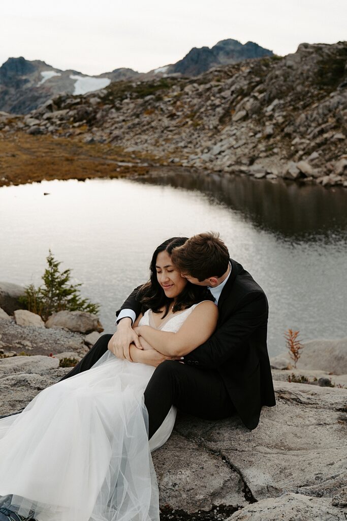 Groom sitting on a rock kissing his bride's cheek while she leans back into his chest.