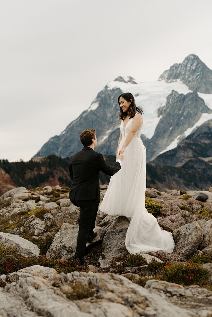 Bride standing on a rock and holding her groom's hand while he climbs the rock with views of Mount Shuksan behind them.