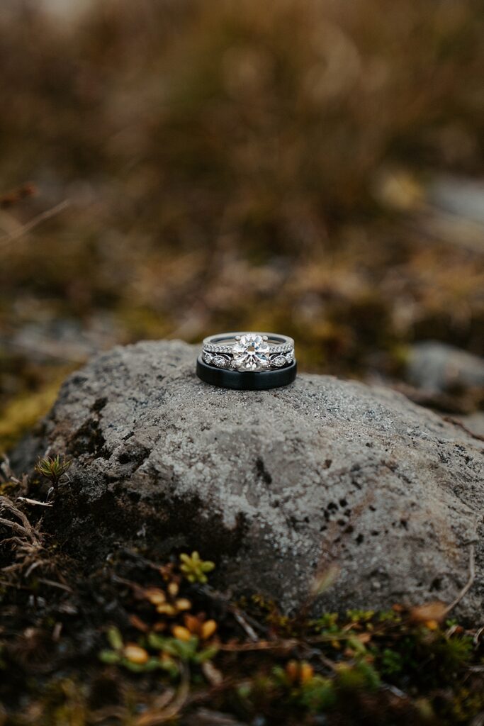 Black wedding band placed on a rock with a wedding band and engagement ring stacked on top.
