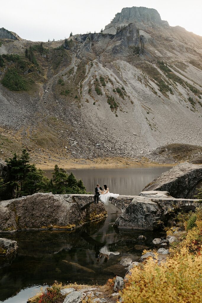 Artist Point elopement photo of bride, groom, and corgi sitting on a stone bridge with a lake and mountain behind them.