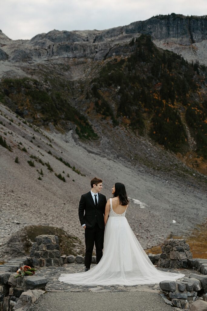 Bride facing away from the camera, but looking at her groom standing next to her with mountains in the background.