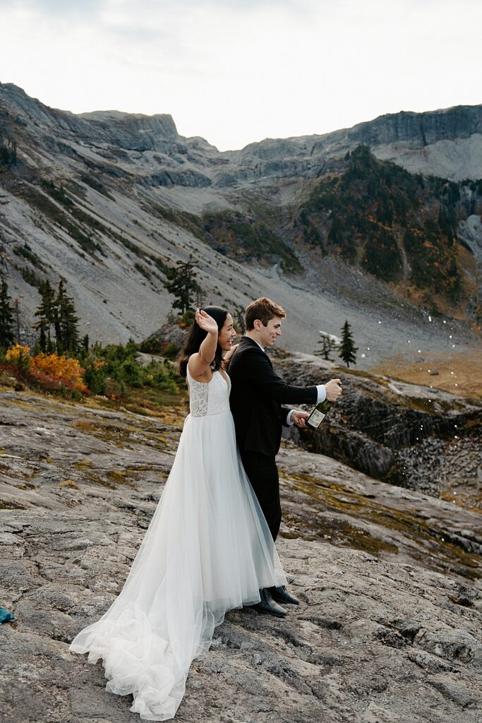 Bride standing behind groom while he pops bubbly to celebrate their Mount Baker elopement.