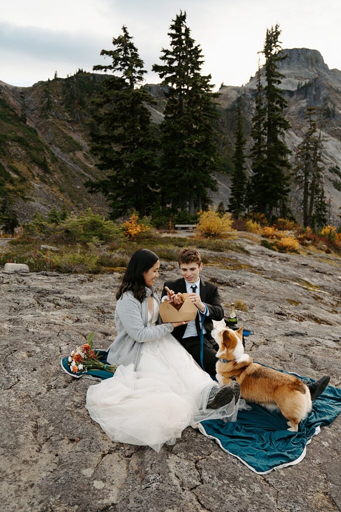 Bride, groom, and corgi pup sitting on a blanket having a picnic to celebrate their Mount Baker-Snoqualmie National Forest elopement.