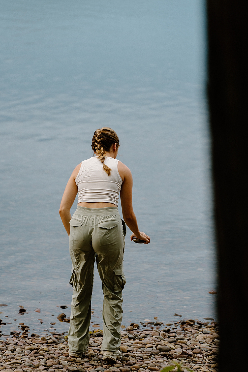 Girl in hiking clothes picking up a rock from the beach for her surprise Glacier Park proposal.