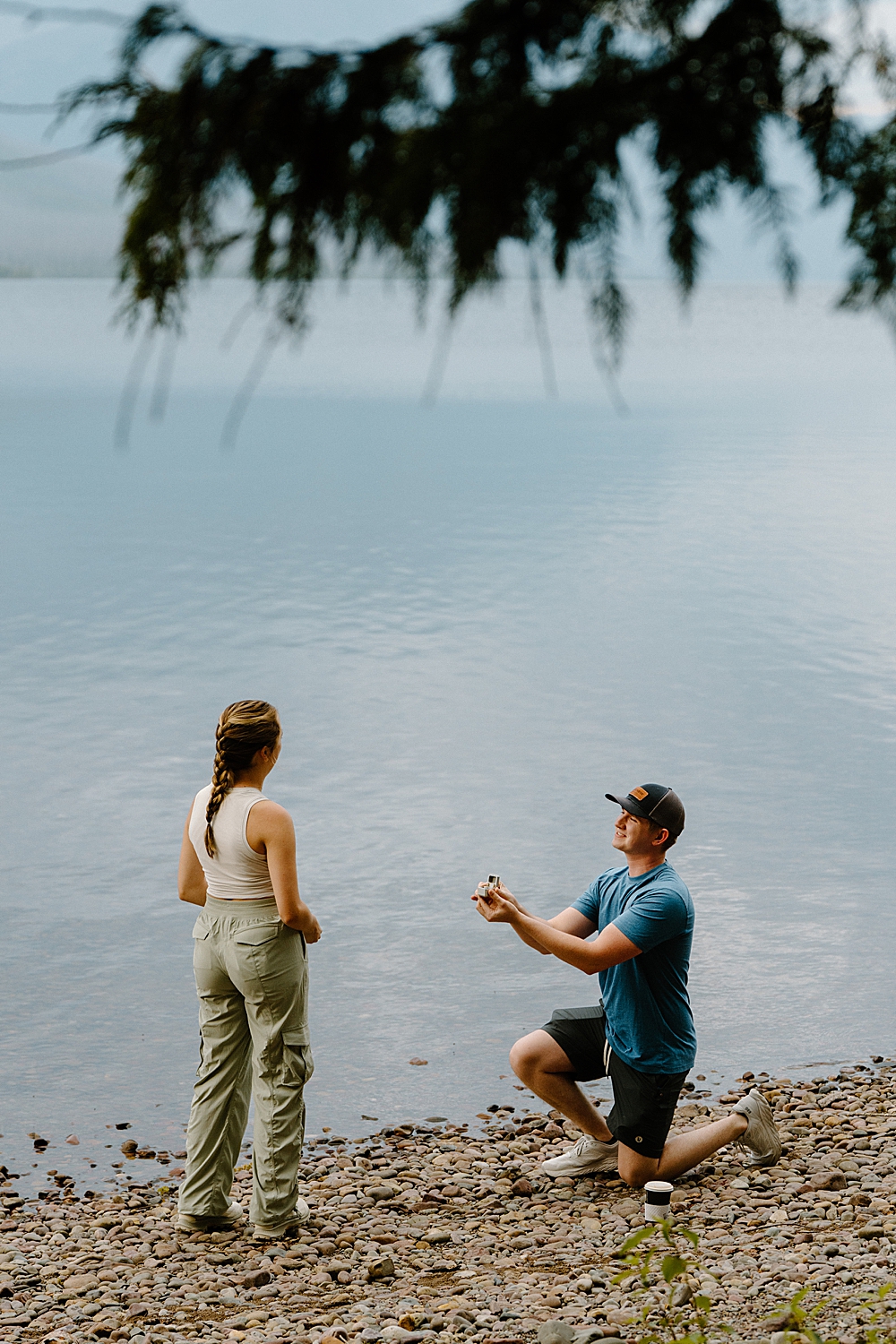 Man in hiking attire proposing to girlfriend on the shore of Lake McDonald in Glacier National Park.