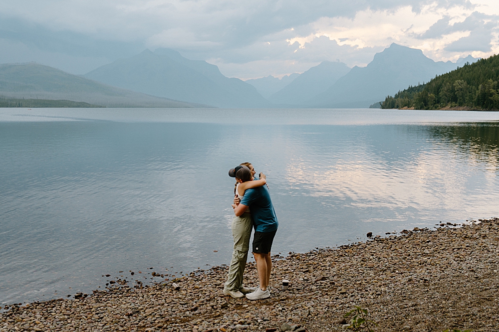 Man and woman in hiking clothes hugging after surprise proposal on Apgar Beach at Lake McDonald in Glacier National Park.