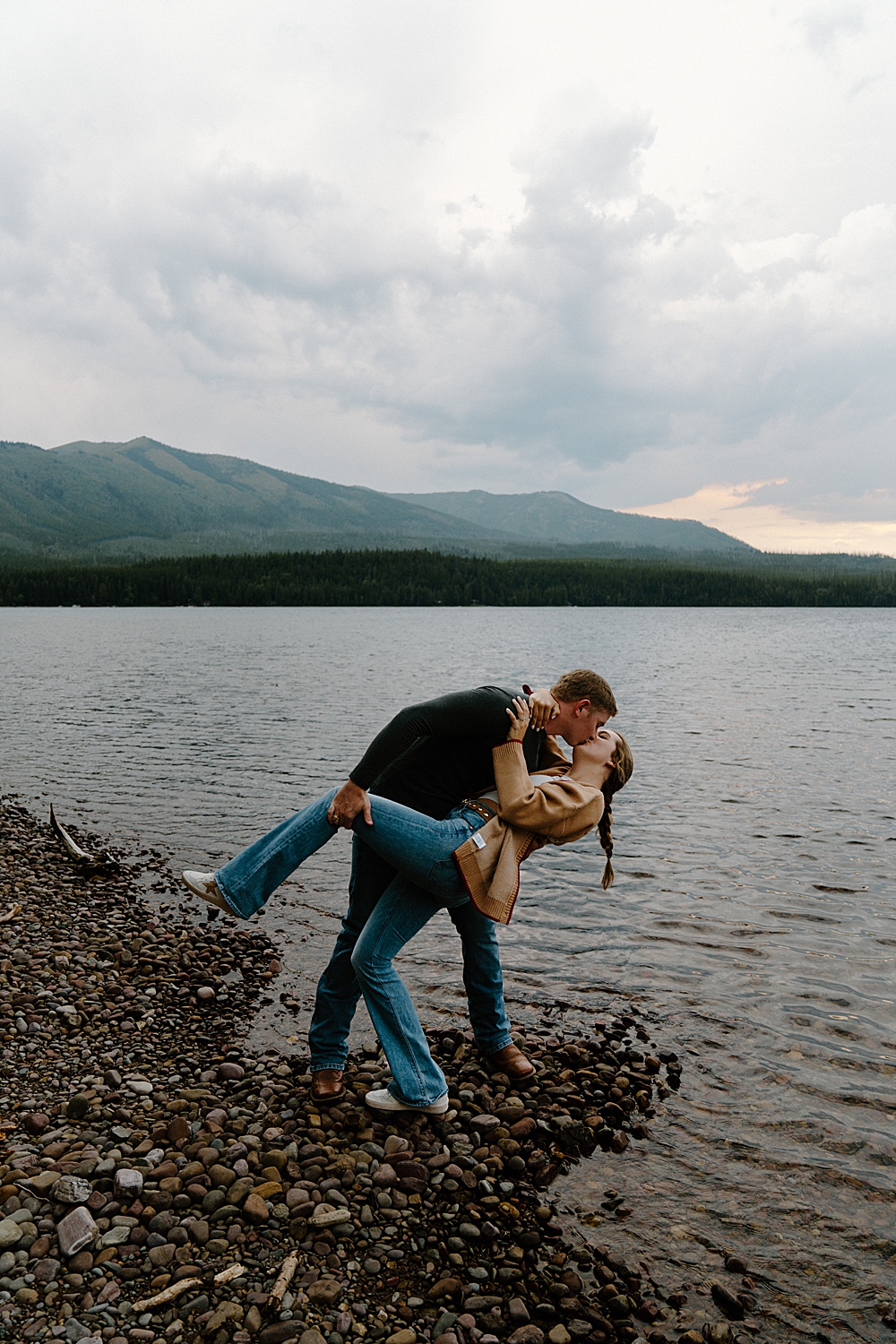Couple doing a dip kiss on the rocky shore of Lake McDonald for their surprise Montana engagement session