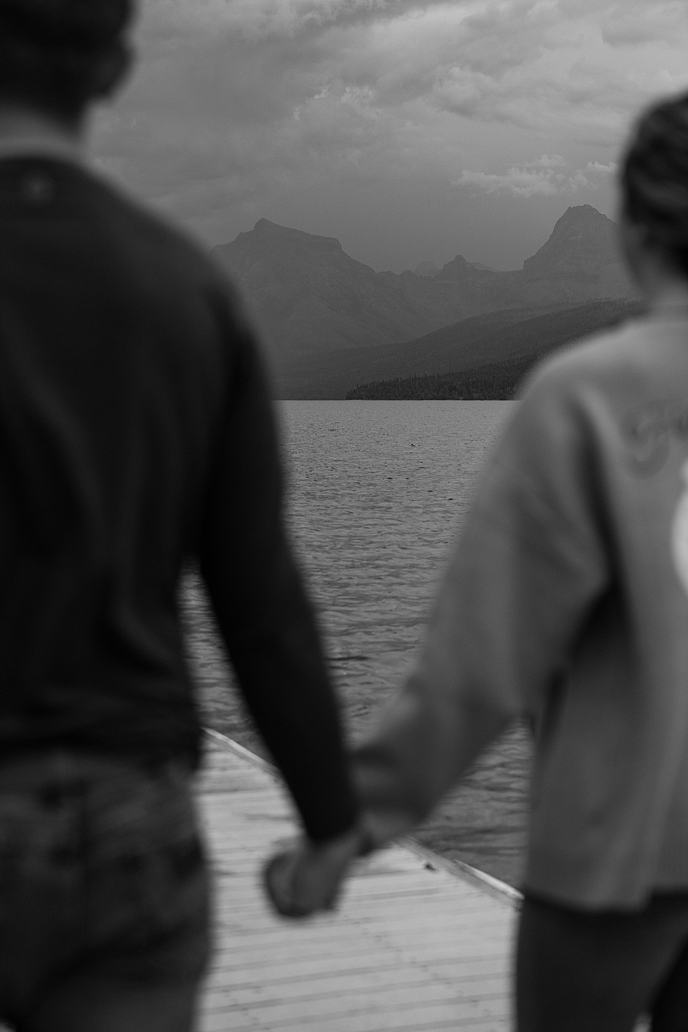 Black & white blurry image of couple holding hands with the mountains at Lake McDonald in focus