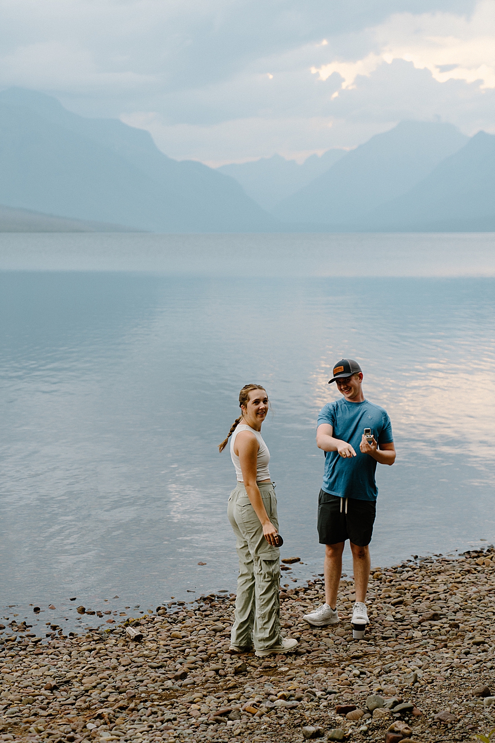Man in hiking attire with a ring box pointing for his girlfriend to notice the photographer's that just captured their surprise Lake McDonald proposal on the beach.