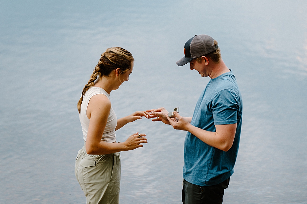 Man in a blue t-shirt and shorts putting an engagement ring on a girl's hand after their surprise Glacier Park proposal.