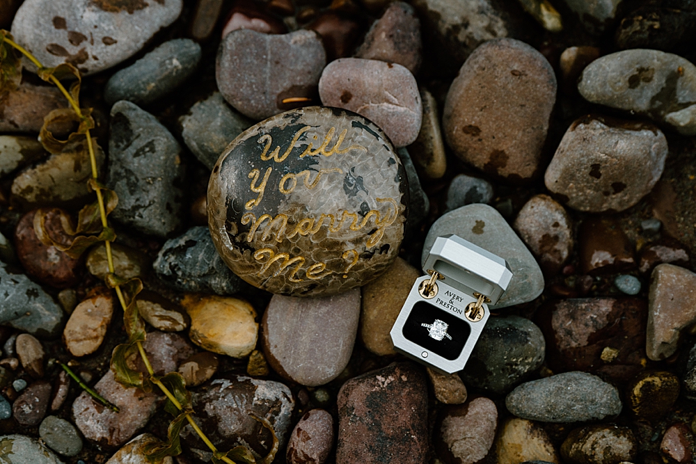 Silver ring box with engagement ring and a rock that reads "Will you marry me?"