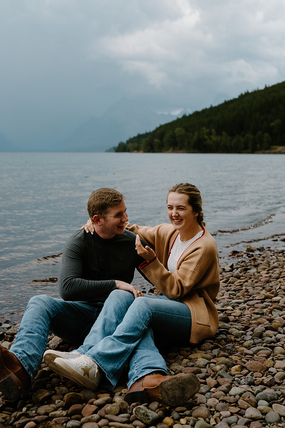 Couple sitting on the beach at Lake McDonald laughing together during their engagement session taken by a local Montana engagement photographer