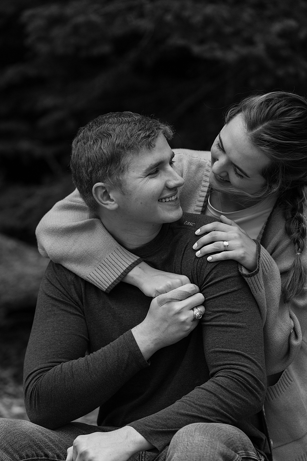 Black & white of girl with her arms around her fiancés shoulders, him holding her hand and looking at her smiling