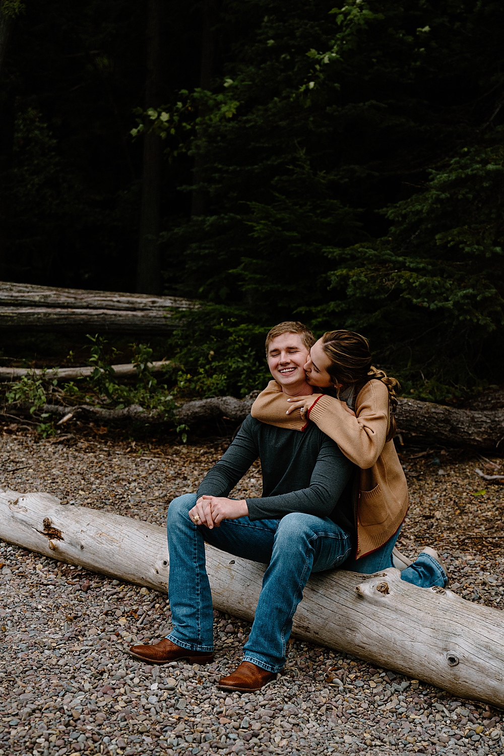 Man sitting on a piece of driftwood with his fiancée kneeling behind him and wrapping her arms around his shoulders while kissing his cheek