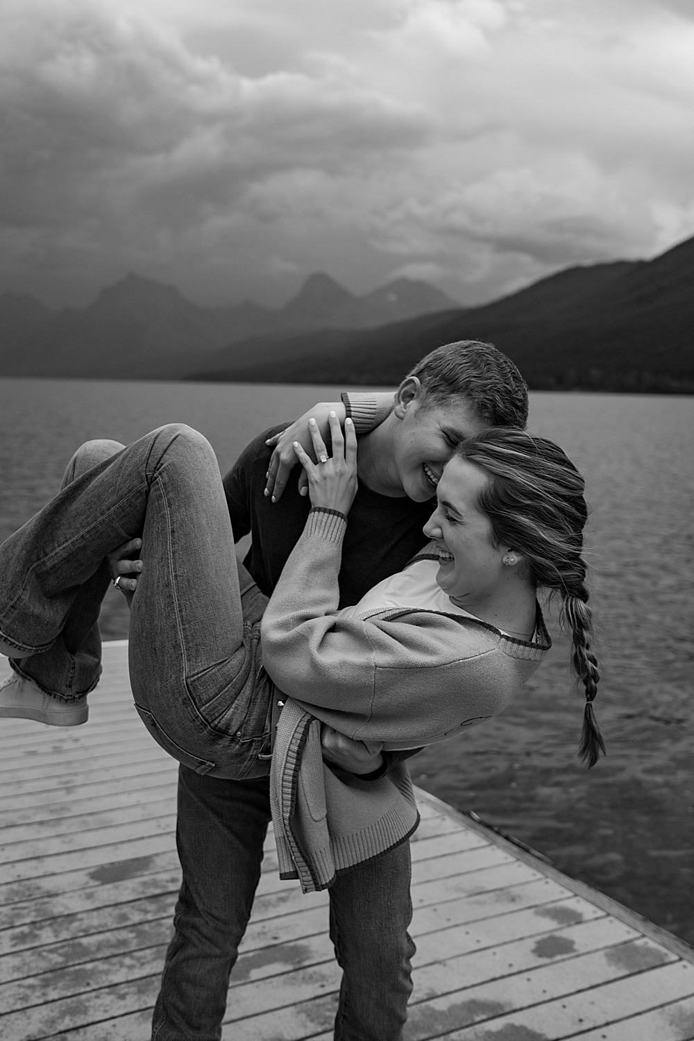 Black & white of man holding his fiancée and tipping her back while they're both smiling and standing on a dock at Lake McDonald