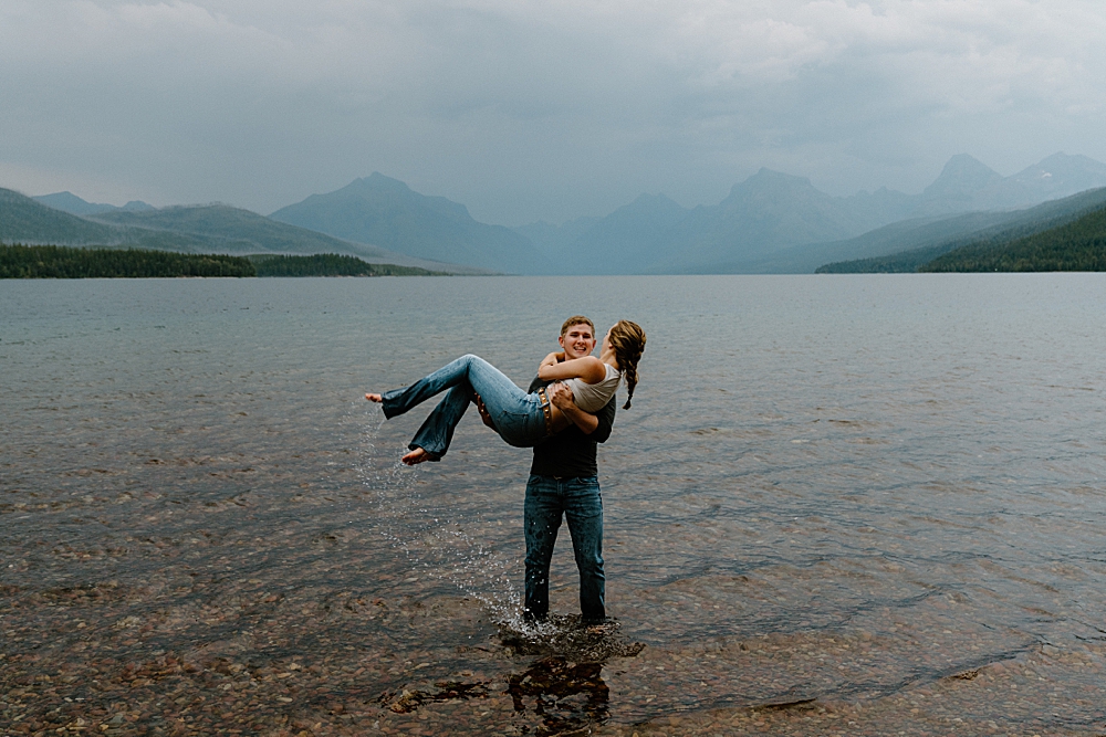 Man holding his girlfriend in his arms while standing in the water at Lake McDonald for their moody/rainy engagement session