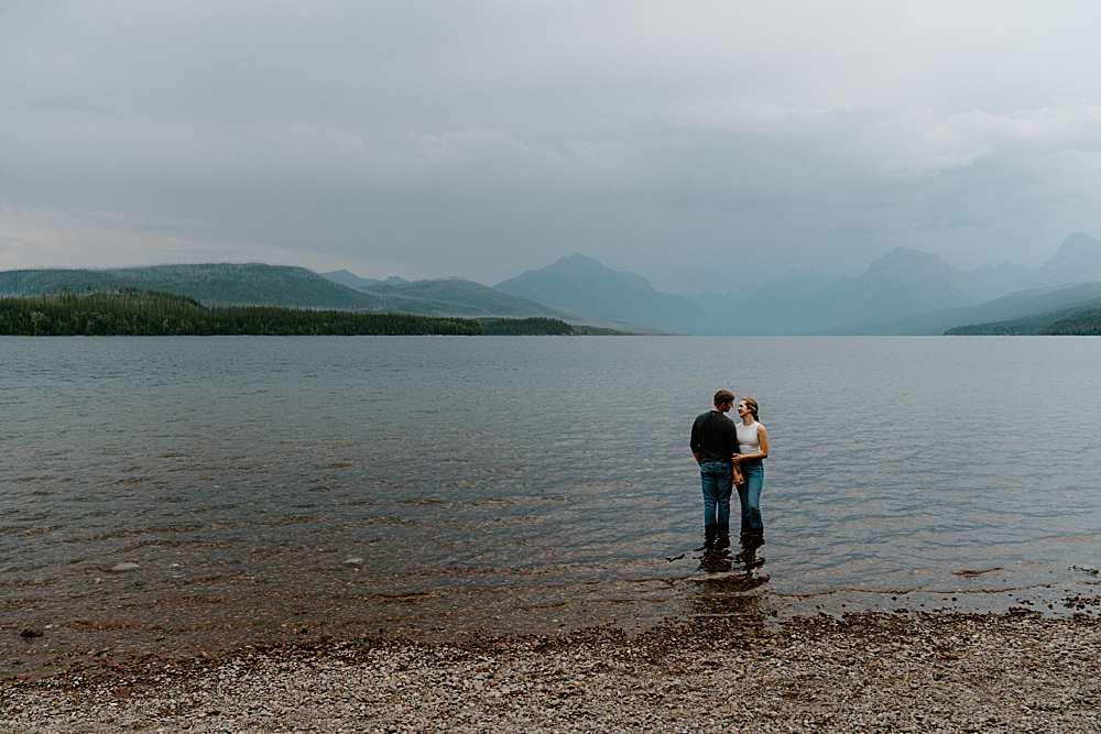 Couple standing in the water at Lake McDonald with a cloudy sky and moody mountains in the background