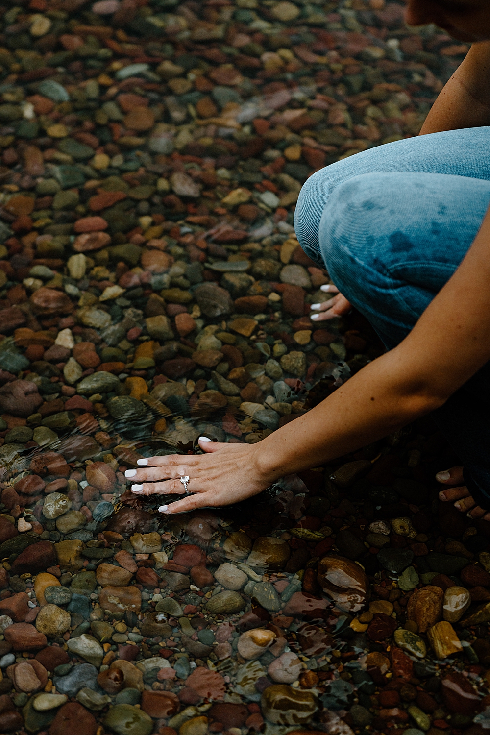 Girl putting her hand with her engagement ring in the water at Lake McDonald touching the colorful rocks