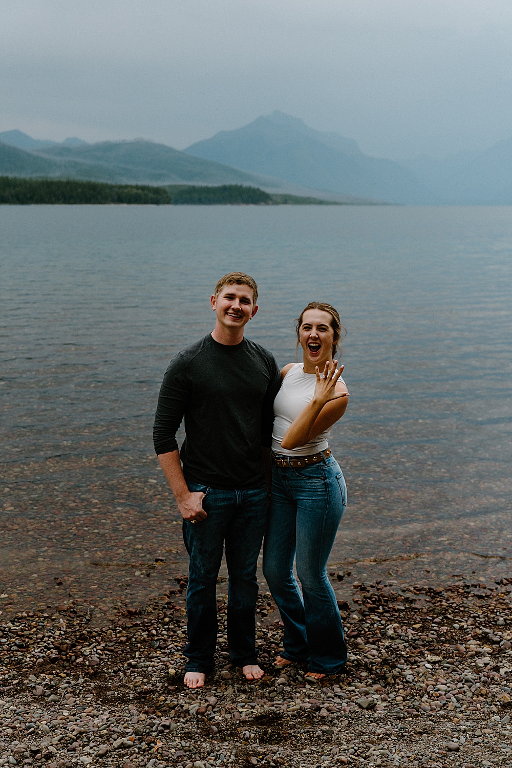 Newly engaged couple standing barefoot at Lake McDonald with the girl showing off her new ring!